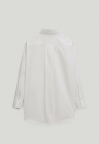 Biała koszula z długim rękawem pokazana z tyłu, z standardowym kołnierzykiem i zapinanymi mankietami na jednolitym jasnym tle.