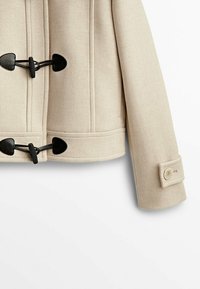 Beżowa kurtka z mieszanki wełny z czarnymi zapięciami na toggles, strukturalnymi szwami i akcentem guzikowym na rękawie, charakteryzująca się czystym designem.