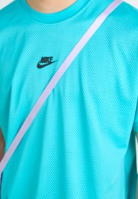 Osoba v tyrkysovém síťovém tričku Nike s diagonálním světle fialovým pásem přes hruď.