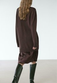 Brązowy, oversize'owy sweter z prążkowanymi mankietami, w połączeniu z ciemnobrązową satynową spódnicą i czarnymi skórzanymi kozakami do kolan, widziany od tyłu.