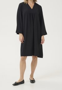 Robe noire longueur genou avec un col en V, des manches longues et une coupe ample. Portée avec des chaussures plates noires. Tissu lisse sans motifs visibles.