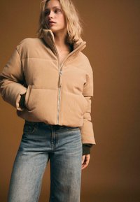 Beżowa kurtka puchowa z korduroju z wysokim kołnierzem, zamkiem błyskawicznym z przodu i ściągaczami, w zestawieniu z niebieskimi dżinsami o wysokim stanie na brązowym tle.