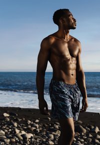 Zwemshorts met een donkerblauwe basis, voorzien van een tropisch bladerenpatroon in groen en rood, met een trekkoord in de taille, gedragen door een naaktbovenlijfs individu.