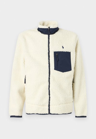 Fleece jacket in cream with a navy zippered chest pocket, high collar, and contrasting trim. Features a textured, soft exterior.