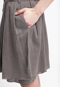 Falda de lino gris con una textura suave, diseño plisado y bolsillo lateral; cuenta con un cierre de botón cerca de la cintura, cayendo justo por encima de la rodilla.