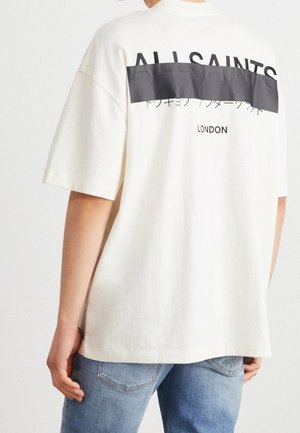 Person wearing white oversized t-shirt with “ALLSAINTS” text partially covered by black bar and “LONDON” printed below, paired with blue jeans.