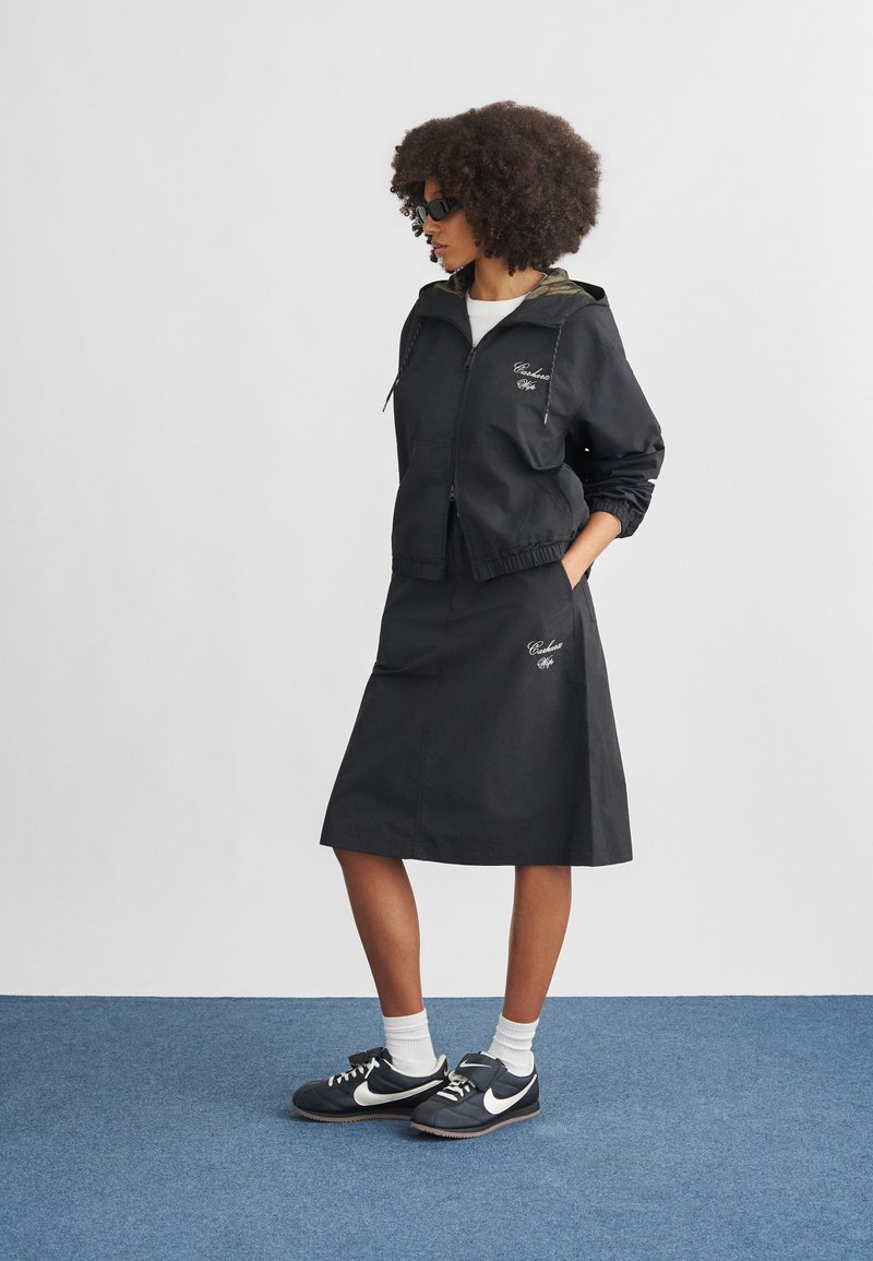 Black windbreaker jacket with an elastic hem, green lining, and embroidered detail, paired with a matching black skirt and black Nike shoes.