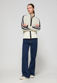 Kabelstickad krämfärgad tröja med hög krage och framdragkedja, med svarta randiga detaljer på ärmarna. Kombinerad med blå utsvängda jeans.