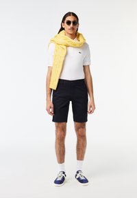 Polo bianco, pantaloni corti blu scuro, sciarpa di lana gialla e sneakers blu con suola bianca. Gli accessori includono occhiali da sole. Il modello è in piedi.