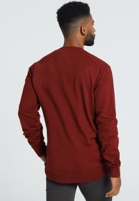 Pull à manches longues de couleur rouge riche, fabriqué en tissu doux. Il possède un col rond et des poignets et une taille côtelés. Texture lisse.