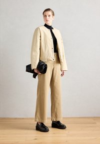 Beige cropped jakke, svart turtleneck, brune vide bukser og svarte plattform loafers. Svart clutch med tekstur i hånden, minimalistisk design.