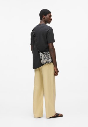 Hombre joven que lleva una camiseta negra, pantalones anchos beige, sandalias negras y un bolso bandolera con estampado en blanco y negro.