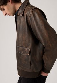 Brown leather jacket with a zip front, large patch pocket, ribbed cuffs, and a slightly distressed finish. Tailored fit and classic collar.