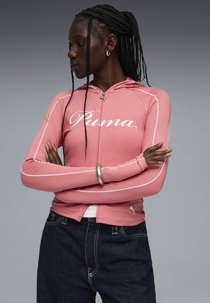 Kvinna med flätat hår som bär en rosa Puma-hoodie med dragkedja och mörka jeans, med armarna korsade, tittar åt vänster mot en grå bakgrund.