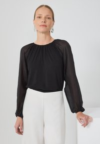 Fekete blúz hosszú, áttetsző ujjal és ráncolt nyakkivágással, fehér nadrággal párosítva. Az anyag simának és könnyűnek tűnik.