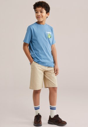 Chlapec s kučeravými vlasmi, oblečený v modrom tričku s potlačou limetiek, béžových šortkách, bielych ponožkách s modrými pruhmi a hnedých teniskách, stojaci s rukami v vreckách.