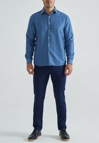 Chemise bleu à boutons avec un petit motif floral blanc, manches longues, col pointu et boutons blancs, associée à un pantalon marine et des chaussures noires.