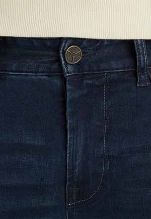 Dark blue denim jeans with a visible button featuring an engraved logo, subtle stitching details, and a smooth texture.