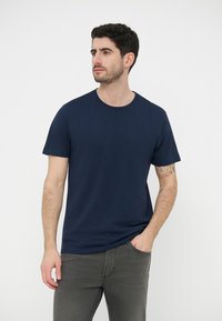 Άνδρας με κοντά σκούρα μαλλιά, φορώντας μπλε σκουρό t-shirt και γκρι τζιν, όρθιος με το ένα χέρι στην τσέπη, πάνω σε λευκό φόντο.