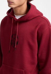 Sudadera color burdeos con bolsillo frontal, decorada con un diseño gráfico negro y cordones con puntas metálicas. Material de mezcla suave de algodón.