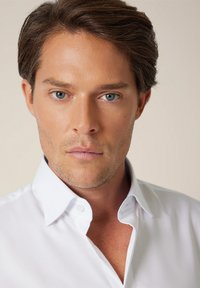 Zbliżenie na mężczyznę noszącego białą koszulę zapinaną na guziki z kołnierzem typu spread. Gładka tkanina, krótkie rękawy i widoczne detale szwów.