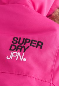 Rozā audums ar izšuvumu "SUPER DRY JPN." melnā un baltā krāsā, ar gludu tekstūru un elegantu apdari.