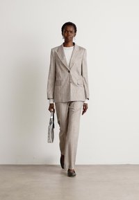 Hellgrauer, strukturierter Anzug mit einem taillierten Blazer und geraden Hosen, kombiniert mit einer weißen Bluse und einer transparenten Handtasche. Schwarze Ballerinas.