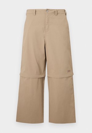 Béžové nastaviteľné nohavice s odnímateľnými dolnými časťami na zips, pútkami na opasok, bočnými vreckami a zapínaním na gombíky, zobrazené na bielom pozadí.