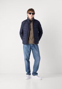 Jūros mėlynos pūkinės striukės su aukštu apykakle, šviesiai rudos spalvos golfas, šviesiai mėlyni džinsai ir balti sportbačiai. Modelis dėvi juodus akinius nuo saulės.