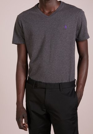 Homme portant un t-shirt à manches courtes col en V gris foncé avec un petit logo violet, assorti à un pantalon noir et une ceinture noire.