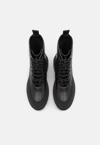 Une paire de bottines en cuir noir à lacets, avec des tirettes, des bouts ronds et des semelles texturées, vues de dessus sur un fond blanc.
