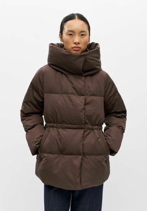 Femme portant une doudoune marron avec un col haut et large, les mains dans les poches latérales, sur un fond clair uni.