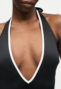 Κοντινό πλάνο ενός ατόμου που φοράει μαύρο τοπ με λαιμόκοψη halter και βαθύ V με λευκή διακόσμηση, σε απλό φόντο.