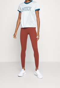 Person med vit Nike T-shirt med blåa detaljer, rostfärgade leggings och vita träningsskor som står mot en enfärgad bakgrund.