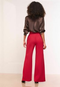 Blusa di raso marrone con maniche arrotolate, abbinata a pantaloni larghi rossi a vita alta, caratterizzati da una texture liscia e un taglio lungo fino al pavimento.