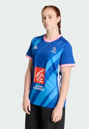 Femme aux cheveux roux tressés portant un maillot de rugby bleu de France avec une bordure rose, un logo de sponsor et un pantalon noir, debout sur un fond uni.