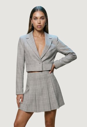 Ung kvinde iført en grå ternet cropped blazer med en enkelt knap og et matchende plisseret miniskørt, stående med den ene hånd på hoften.