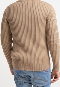 Suéter beige acanalado de manga larga, con un diseño ajustado. Textura de punto visible y detalle de cuello redondo, combinado con jeans azules.