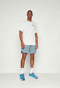 Mann som står iført hvit t-skjorte, lyseblå shorts, hvite Nike-sokker og blå joggesko mot en enkel lys bakgrunn.