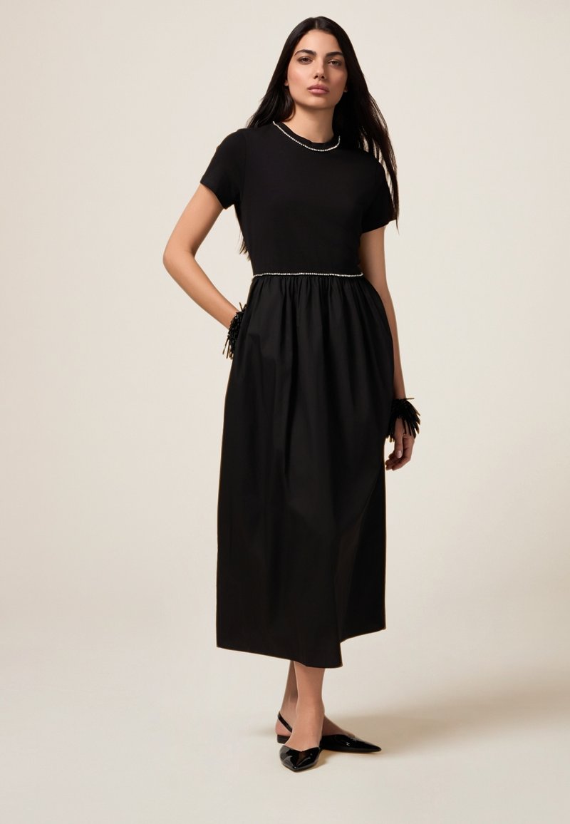 Schwarzes Kleid mit tailliertem Oberteil und gerafftem Rock, mit einem perlenähnlichen Ausschnitt. Getragen mit eleganten schwarzen Ballerinas und einem Akzent am Handgelenk.