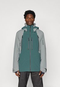 Férfi teal és szürke kültéri dzsekiben, ami több cipzáras zsebből áll, fekete nadrágban, egy sima fehér háttér előtt áll.