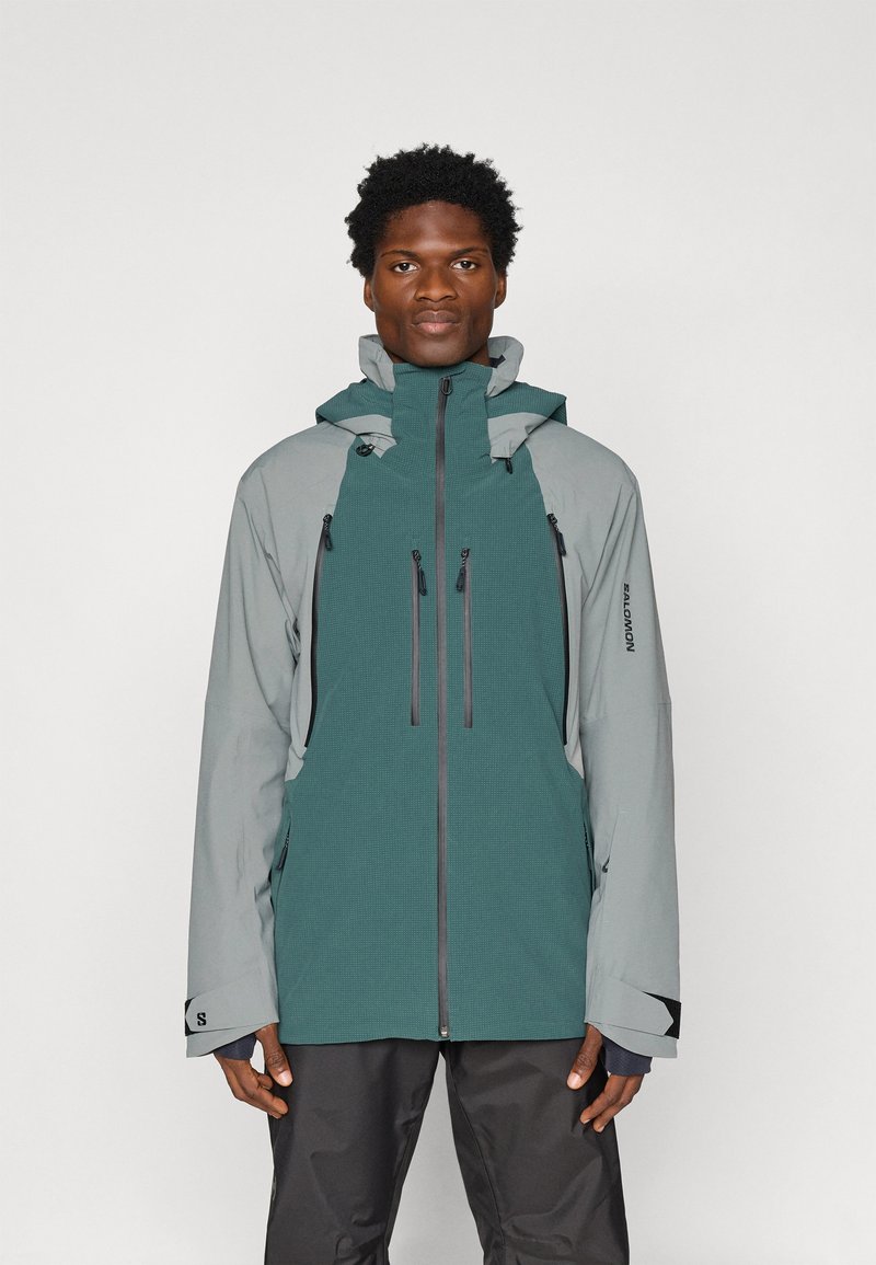 Férfi teal és szürke kültéri dzsekiben, ami több cipzáras zsebből áll, fekete nadrágban, egy sima fehér háttér előtt áll.