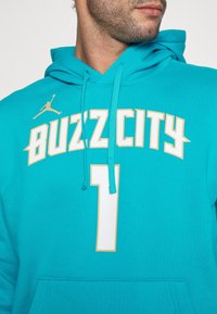 Felpa teal con cappuccio e coulisse, con la scritta "BUZZ CITY" e il numero "1" in bianco e oro. Include un piccolo logo Jordan sulla spalla.