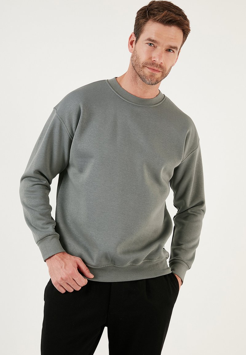 Sudadera gris texturizada con cuello redondo y mangas largas. Corte holgado con puños y dobladillo acanalados. Llevada con pantalones negros.