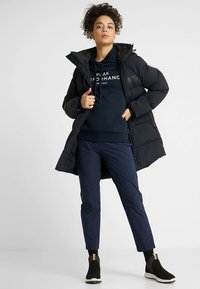 Juoda pūkinė striukė dėvima virš tamsiai mėlynos gobtuvinės su „Peak Performance“ logotipu, derinama su tamsiai mėlynomis siaurėjančiomis kelnėmis ir juodais kulkšnies ilgio batais su baltais padais.