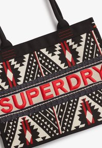 Canvas tas met geometrisch patroon in zwart, crème en rood, met een opvallende rode "SUPERDRY" tekst in het midden.