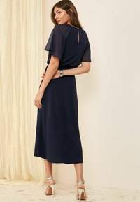 Marineblaues Kleid mit kurzen, durchsichtigen Ärmeln und einem Schlüsselloch-Design am Rücken. Der Rock endet auf Kniehöhe. Getragen mit silbernen Riemchenheels.