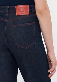 Nærbilde av mørkeblå jeans med røde sømmer og et rødt lærmerke med en rose og merkevarenavnet "ONLY" på livlinjen.