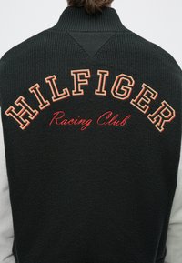 Σκούρο πλεκτό πουλόβερ στην πλάτη με την επιγραφή «HILFIGER» σε μπεζ κεφαλαία γράμματα και «Racing Club» κεντημένα με κόκκινα γράμματα σε καλλιγραφικό στυλ στο πάνω μέρος της πλάτης.