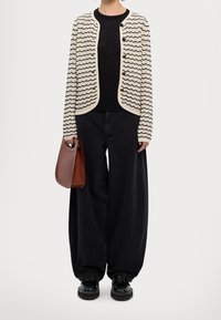 Neulettu kerma-valkoinen cardigan, jossa on aaltoilevat mustat raidat ja napit mustan neuleen päällä; yhdistetty suurikokoisiin mustiin housuihin ja ruskeaan laukkuun.